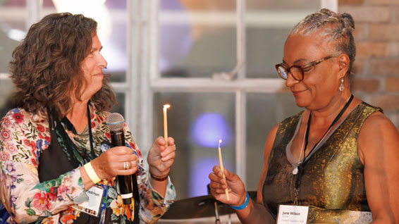 Two women participate in a candle-lighting ceremony indoors. One woman, Ellen Friedman, holding a microphone and a lit candle, lights the candle of the other woman, who is wearing glasses and a name tag that reads 'June Wilson.'