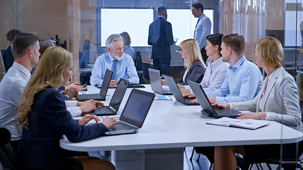 Corporate meeting scene showing a group of business professionals seated around a white conference table in a glass-walled office. Most are using laptops, and one older man appears to be leading the discussion. Two additional people stand conversing in the background, silhouetted against large windows. The setting feels formal and hierarchical.
