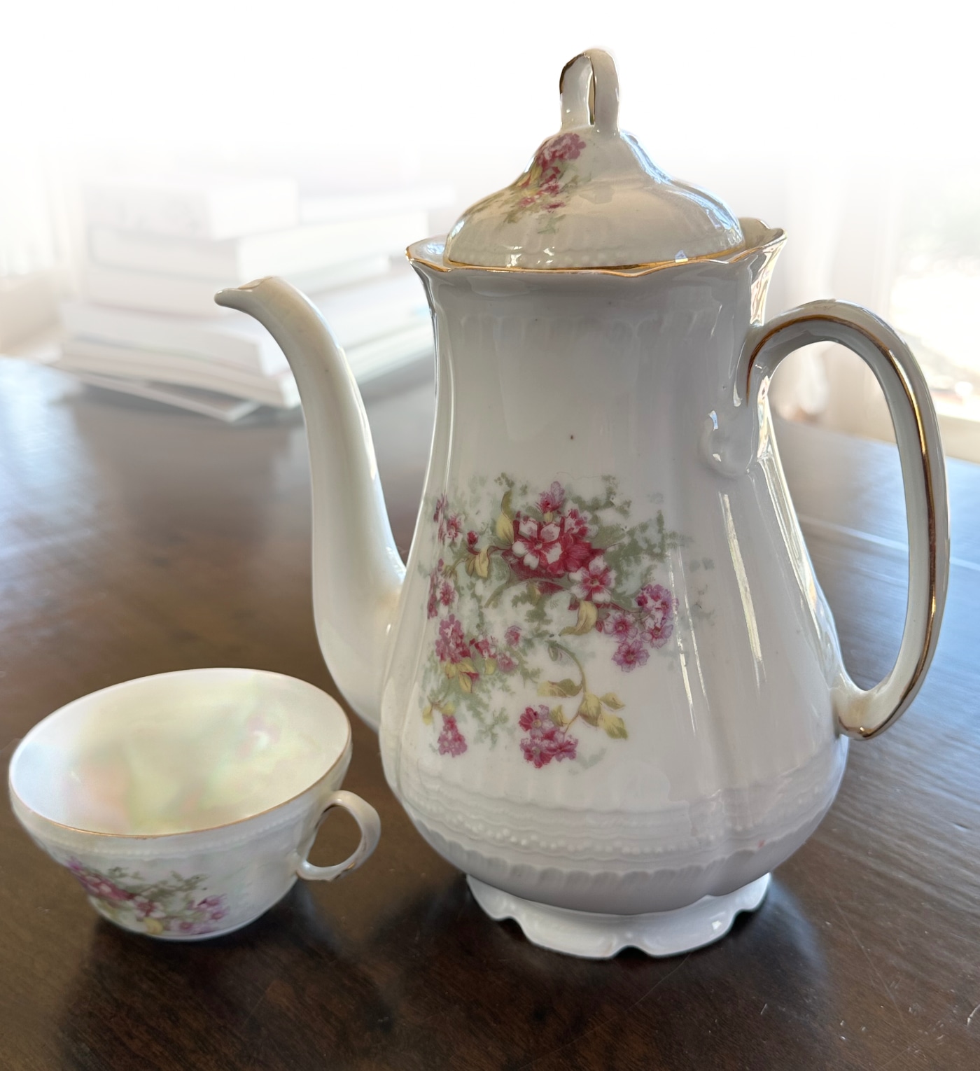 On a brown wooden table sits a tall white porcelain teapot with a lid and long spout. The teapot is decorated on the side with a small cluster of dark red flowers, and gilded on the rim and along the handle in gold. Next to it is a teacup with similar decorations. The background fades to white in the top half of the photo, making the teacup and teapot stand out from the background as if emerging from the photograph.