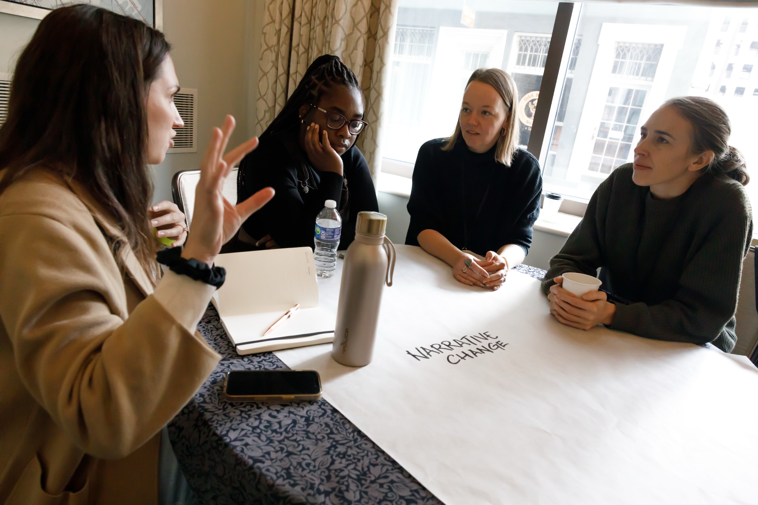 Four women sit around a small table in a hotel meeting room. A large sheet of white paper is spread over the table. The paper is blank except for the words "Narrative Change" in large black capital letters in the center. The woman on the left is gesturing as she speaks, and the other three women are listening.