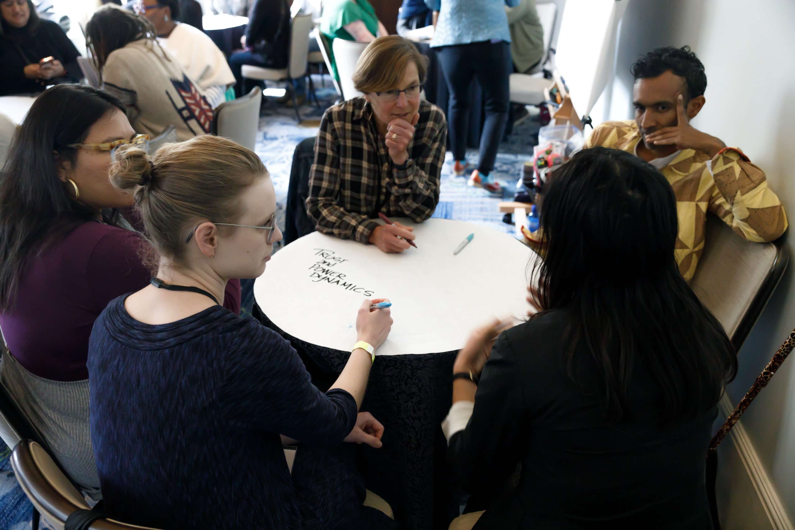 Five people--four women and one man--sit around a small round table in a hotel conference room. Behind them, partially cropped and out of focus, are other groups sitting around similar tables. The table is covered entirely in a sheet of round white paper, which is blank except for the words "Trust and Power Dynamics" hand written in decorative capital letters. In the foreground one of the women has her back to the camera and is gesturing as she speaks to the group. The other four are looking at her and listening.